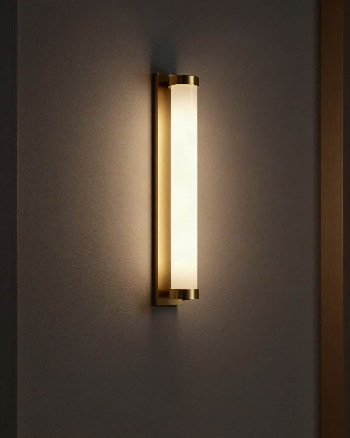 A modern wall sconce, a slim brass bar with a frosted glass cylinder glowing warm on a dark plaster wall