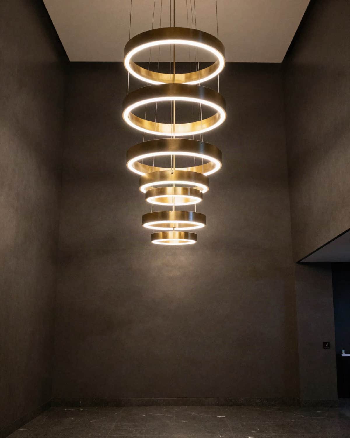 A modern chandelier of tiered brass rings with warm LED bands, hanging in a double-height hall with dark plaster walls