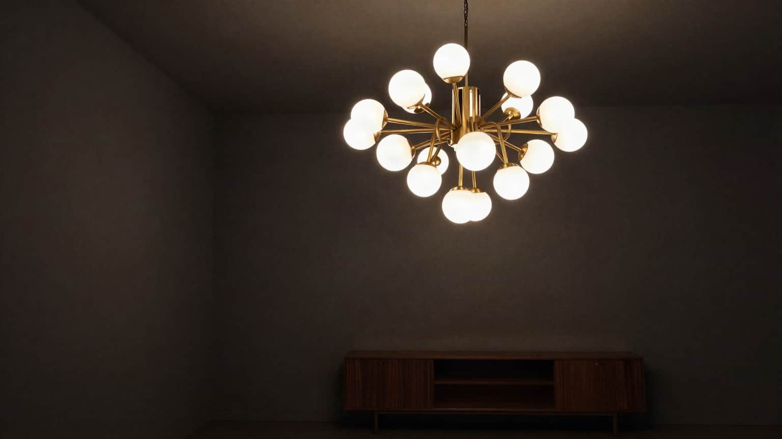 A modern brass chandelier of frosted glass globes hanging in a dark contemporary living room, lit warm